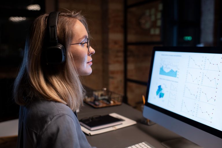 ulher com fones de ouvido analisando gráficos e dados em uma tela de computador em um escritório.