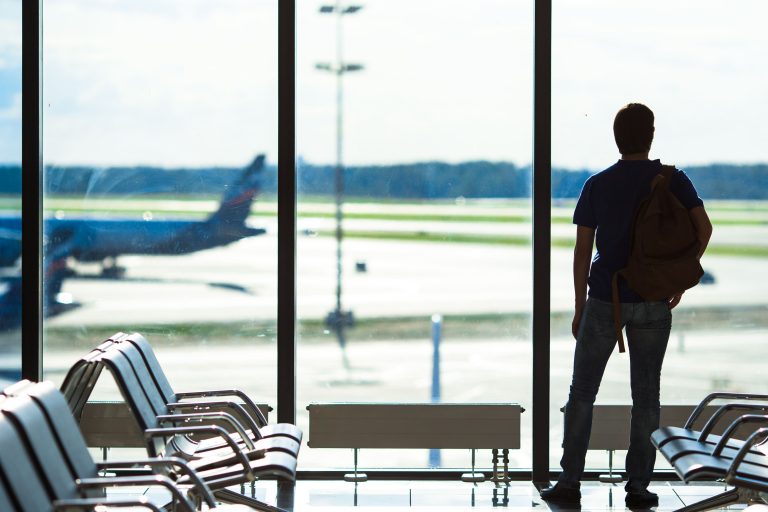 Homem esperando no aeroporto com vista para a pista e avião ao fundo.